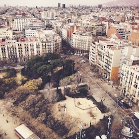 An aerial view of an urban neighborhood featuring a mix of residential and commercial buildings. The streets are lined with trees, and a park with trees and pathways is visible in the foreground. The buildings are mostly multi-story, displaying a variety of architectural styles. The atmosphere appears calm and the city extends to the horizon.