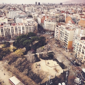 An aerial view of an urban neighborhood featuring a mix of residential and commercial buildings. The streets are lined with trees, and a park with trees and pathways is visible in the foreground. The buildings are mostly multi-story, displaying a variety of architectural styles. The atmosphere appears calm and the city extends to the horizon.
