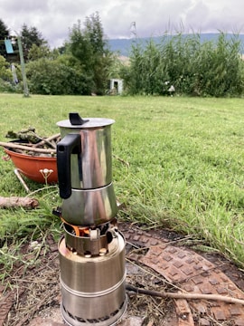A metal stovetop coffee maker sits atop a small portable camping stove, with visible flames beneath it. The setup is on a patch of grass with pieces of wood and a rusty manhole cover nearby. A lush garden with tall grasses and trees forms the background under a cloudy sky.