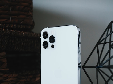 A modern smartphone with a sleek white finish and multiple camera lenses is prominently positioned in front of a woven basket and a geometric metal structure. The lighting and composition emphasize the device's contemporary design.
