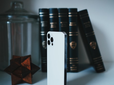 A sleek, white smartphone with a prominent camera setup stands upright on a surface. In the background, there are several black leather-bound books with gold accents, arranged neatly in an upright position. To the left, a geometric wooden puzzle and a large glass jar add an artistic touch to the scene.