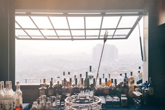 A collection of various liquor bottles and barware arranged neatly on a bar counter, with a panoramic view of a city skyline visible through a large window in the background. The cityscape appears hazy, likely due to fog or smog, and high-rise buildings can be seen in the distance. A long cocktail stirring spoon is prominently placed in the center, adding a focus to the bar tools and setting.