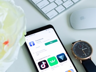 A smartphone displaying the Facebook app page on a light-colored table. Nearby, there is a gold-colored wristwatch with a black strap, a white Apple keyboard, and a white Apple Magic Mouse. A blurred white flower is also visible in the foreground.