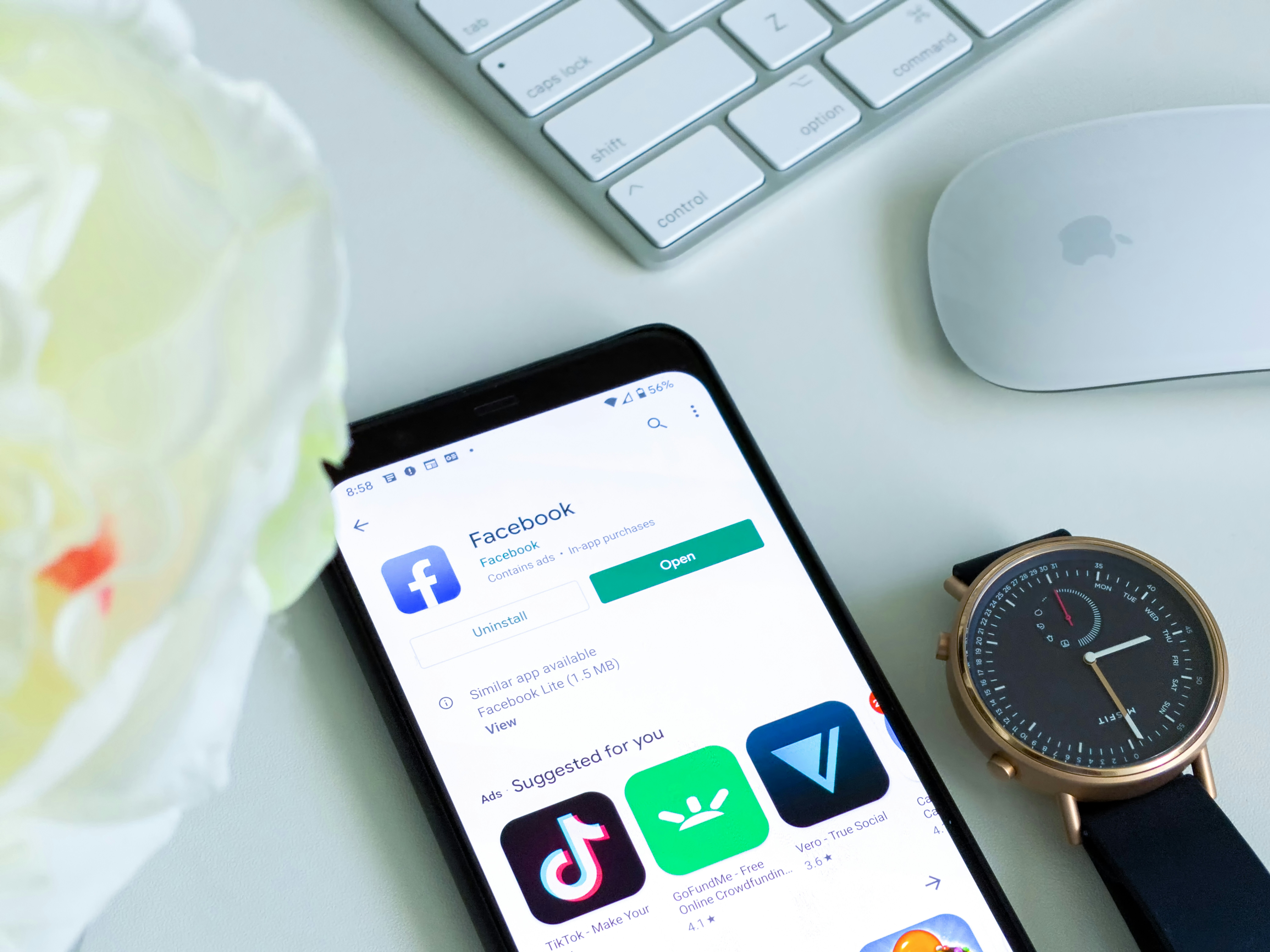 A phone showing the Facebook app page and a Misfit Smartwatch on a home office workstation, next to an apple mouse and keyboard.