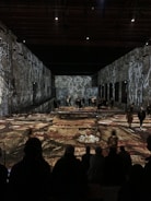 A large, darkened room with visual art projections on the walls and floor, featuring abstract figures and patterns. People are scattered throughout the space, both standing and sitting, observing the artistic display.