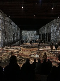 A large, darkened room with visual art projections on the walls and floor, featuring abstract figures and patterns. People are scattered throughout the space, both standing and sitting, observing the artistic display.
