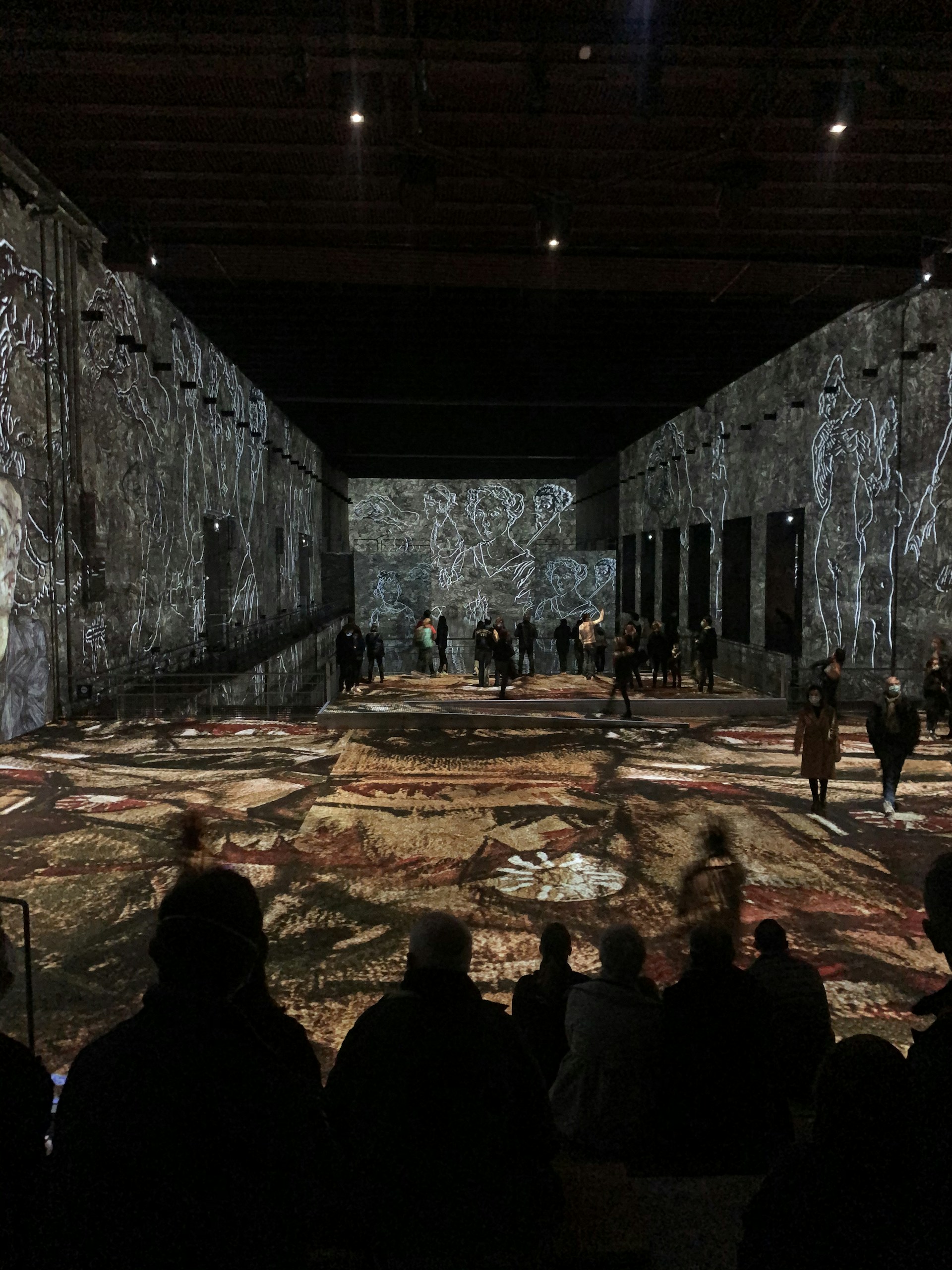 A large, darkened room with visual art projections on the walls and floor, featuring abstract figures and patterns. People are scattered throughout the space, both standing and sitting, observing the artistic display.