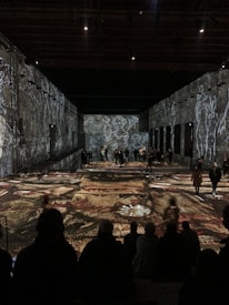 A large, darkened room with visual art projections on the walls and floor, featuring abstract figures and patterns. People are scattered throughout the space, both standing and sitting, observing the artistic display.
