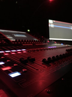 A close-up of a sound mixing console illuminated by red and dim blue lights. The scene is dark, with the interface showing various buttons, sliders, and screens displaying graphs or waveforms. The equipment suggests a professional audio setup, possibly in a recording studio or live music venue.