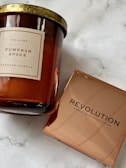 A scented candle with a label indicating 'Pumpkin Spice' from DW Home alongside a makeup product from Revolution Makeup Revolution London. The candle has a golden lid and an amber-colored glass container. The Revolution product is packaged in a reflective, copper-hued box.