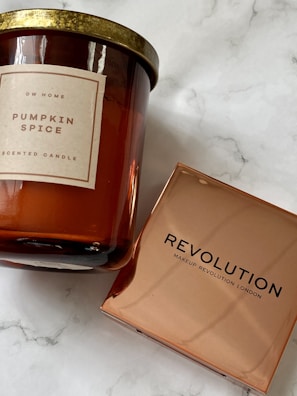 A scented candle with a label indicating 'Pumpkin Spice' from DW Home alongside a makeup product from Revolution Makeup Revolution London. The candle has a golden lid and an amber-colored glass container. The Revolution product is packaged in a reflective, copper-hued box.