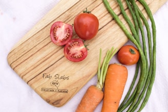 A wooden cutting board with the brand 'Fab Slabs' has various fresh vegetables arranged on it, including sliced and whole tomatoes, carrots with green tops, and several long green beans. The board is set on a white cloth background.