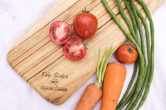 A wooden cutting board with the brand 'Fab Slabs' has various fresh vegetables arranged on it, including sliced and whole tomatoes, carrots with green tops, and several long green beans. The board is set on a white cloth background.