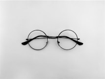A pair of round, black-framed eyeglasses placed on a plain white background. The symmetrical design and minimalist setting create a simple yet elegant aesthetic.