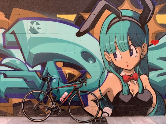 A vibrant street art mural featuring an anime-style character with teal hair and bunny ears, wearing a red bow tie and a black outfit. In front of the mural, a black bicycle with a pink water bottle is parked on the sidewalk.
