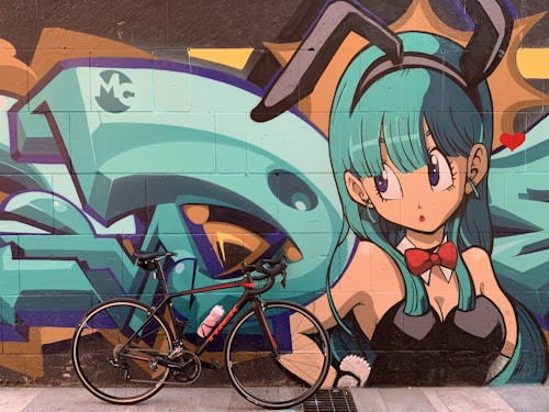 A vibrant street art mural featuring an anime-style character with teal hair and bunny ears, wearing a red bow tie and a black outfit. In front of the mural, a black bicycle with a pink water bottle is parked on the sidewalk.