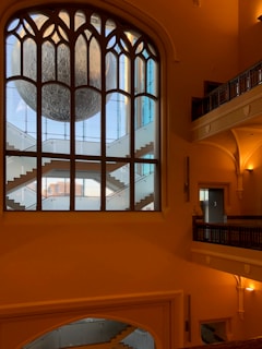 A large, ornate interior window frames an art installation resembling a detailed lunar surface, suspended behind a modern, geometric staircase within a building that features classic architectural elements such as arches and decorative railings.