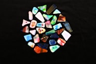 A collection of colorful, uniquely shaped glass pieces arranged in a circular pattern on a black background. Each piece is distinct, featuring various colors and designs, including swirls, speckles, and layered textures.