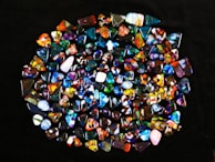 A collection of colorful, polished gemstones scattered on a dark surface. The stones have a variety of shapes, including triangles and ovals, with complex patterns and vibrant hues like blues, reds, yellows, and greens.