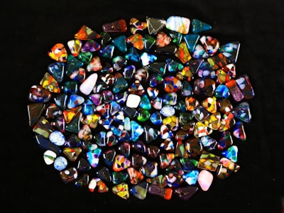 A collection of colorful, polished gemstones scattered on a dark surface. The stones have a variety of shapes, including triangles and ovals, with complex patterns and vibrant hues like blues, reds, yellows, and greens.