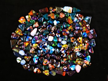 A collection of colorful, polished gemstones scattered on a dark surface. The stones have a variety of shapes, including triangles and ovals, with complex patterns and vibrant hues like blues, reds, yellows, and greens.