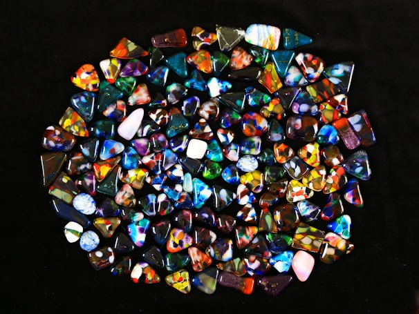 A collection of colorful, polished gemstones scattered on a dark surface. The stones have a variety of shapes, including triangles and ovals, with complex patterns and vibrant hues like blues, reds, yellows, and greens.