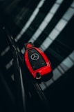 A red car key with a luxury brand logo is resting on a reflective dark surface, likely a windshield. The key features multiple buttons for locking, unlocking, and opening the trunk.