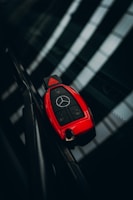 A red car key with a luxury brand logo is resting on a reflective dark surface, likely a windshield. The key features multiple buttons for locking, unlocking, and opening the trunk.
