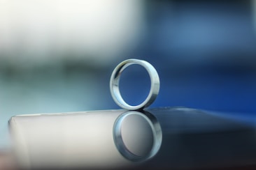 A metallic ring is balanced on its side on a smooth, reflective surface. The background is an unfocused gradient of blues and grays, creating a calm and serene ambiance. The ring casts a clear reflection on the surface beneath it.