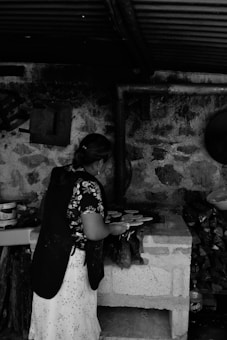 A woman stands by a rustic stone stove, making food in what appears to be a traditional kitchen setting. The surroundings include stacks of firewood, a stone wall, and a corrugated metal roof. The woman is wearing a floral top and a white skirt with an apron, and she is using a spatula to cook.