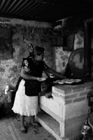 A woman is preparing food on a traditional brick stove in a rustic setting. She is wearing a patterned blouse and apron, focusing on arranging items on a cooking surface. The area has a rough, stone wall and various cooking utensils are visible.