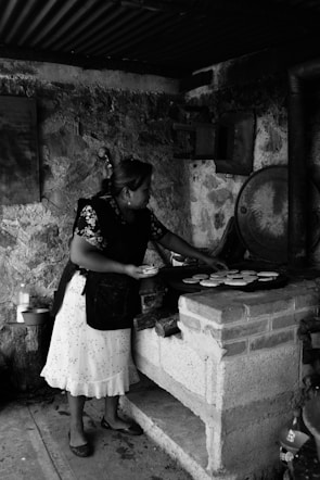 A woman is preparing food on a traditional brick stove in a rustic setting. She is wearing a patterned blouse and apron, focusing on arranging items on a cooking surface. The area has a rough, stone wall and various cooking utensils are visible.