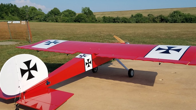 A model airplane with a red body and wings, featuring black and white crosses, sits on a concrete surface in an open grassy field. The plane has a wooden propeller and is positioned near some shrubs and trees, with a clear blue sky above.