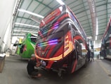 A row of buses with vibrant paint jobs are inside a large garage, undergoing maintenance or customization. One person, wearing a cap and seated on the ground, is working on the rear end of a bus with striking red and black graphics. Another person is standing beside a green bus with an open rear compartment. The ceiling of the garage is metal and has ample overhead lighting, indicating a spacious indoor facility.