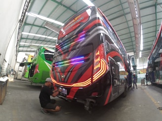 A row of buses with vibrant paint jobs are inside a large garage, undergoing maintenance or customization. One person, wearing a cap and seated on the ground, is working on the rear end of a bus with striking red and black graphics. Another person is standing beside a green bus with an open rear compartment. The ceiling of the garage is metal and has ample overhead lighting, indicating a spacious indoor facility.