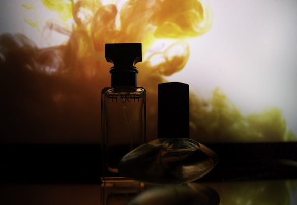 Two perfume bottles are silhouetted against a vibrant, swirling background of yellow and orange hues. The lighting casts a dramatic effect, highlighting the contours and elegance of the glass bottles.