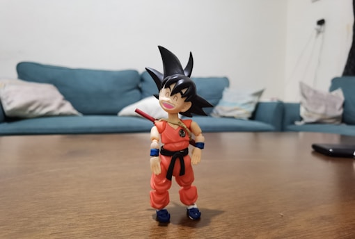 A toy figure of an animated character stands on a wooden table. The figure wears an orange outfit with a blue wristband and features spiky black hair, smiling joyfully. In the background, there is a teal sofa and a few cushions placed on it in a casual setting.