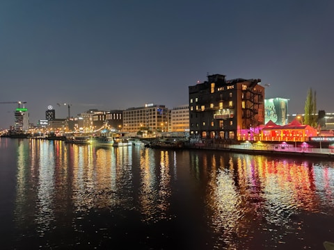 A vibrant urban night scene along a river, featuring illuminated buildings reflecting colorful lights on the water. The landscape includes modern architecture, visible cranes, and signage from various businesses. A lively atmosphere is conveyed with bustling activity and bright neon colors.