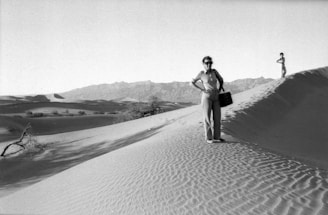 A black and white photograph features two people standing on sand dunes in a desert landscape. One person stands closer to the camera, carrying a handbag, wearing sunglasses and casual clothes. The other person is further away, standing atop another dune. The vast desert stretches into the distance with sparse vegetation and a range of mountains in the background. The sunlight casts long shadows over the rippled patterns in the sand.