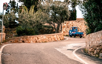 A small blue vehicle drives along a winding road lined with stone walls and surrounded by lush greenery, including olive trees. A traffic sign is visible on the left. The scene suggests a peaceful, rural setting.