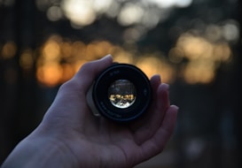 A hand is holding a camera lens, which reflects a scene of trees and the sky. The background is blurred with bokeh lights, creating a warm, soft focus effect as the sun sets.