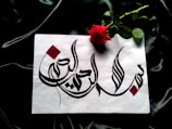 A piece of white paper features intricate Arabic calligraphy in bold black ink, adorned with red geometric accents. Beside the paper lies a vibrant red rose with lush green leaves, all set against a dark, silky fabric background.