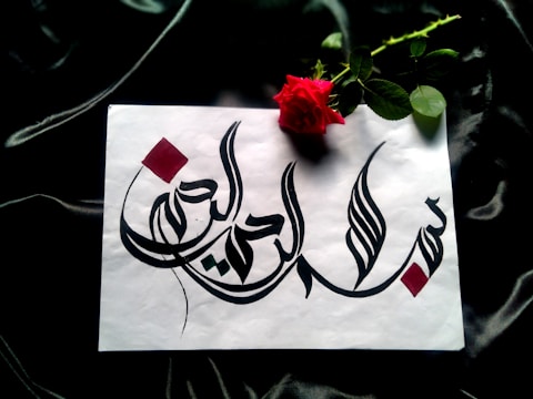 A piece of white paper features intricate Arabic calligraphy in bold black ink, adorned with red geometric accents. Beside the paper lies a vibrant red rose with lush green leaves, all set against a dark, silky fabric background.