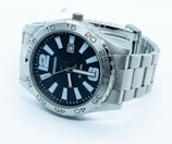 A silver wristwatch with a metal strap and a black dial. The watch face features bold white hour markers and a date display. The bezel is marked with minute intervals and the watch hands are silver-toned. The brand name is visible on the face.