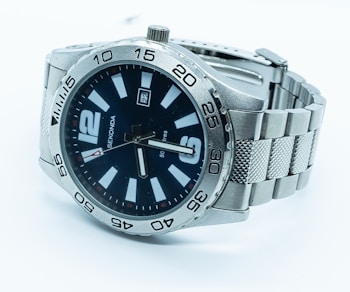 A silver wristwatch with a metal strap and a black dial. The watch face features bold white hour markers and a date display. The bezel is marked with minute intervals and the watch hands are silver-toned. The brand name is visible on the face.