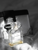 A bottle of Chanel No. 5 Eau de Parfum is placed in the foreground, exhibiting its classic and elegant glass design. Behind it, a Prada Milano bag can be seen, adding a touch of luxury. The background is softly blurred, giving prominence to the perfume bottle.