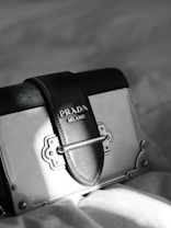 A luxury designer handbag is prominently displayed, featuring a stylish rectangular shape with a sophisticated metal clasp. The bag has a black and gray color scheme with fine stitching details. A Prada Milano logo is elegantly embossed on the leather.