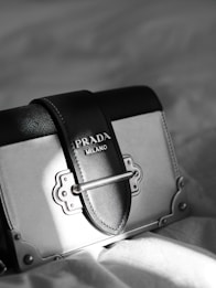 A luxury designer handbag is prominently displayed, featuring a stylish rectangular shape with a sophisticated metal clasp. The bag has a black and gray color scheme with fine stitching details. A Prada Milano logo is elegantly embossed on the leather.