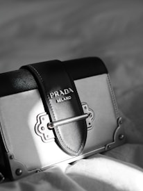A luxury designer handbag is prominently displayed, featuring a stylish rectangular shape with a sophisticated metal clasp. The bag has a black and gray color scheme with fine stitching details. A Prada Milano logo is elegantly embossed on the leather.