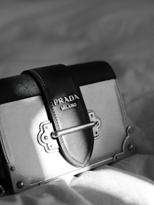 A luxury designer handbag is prominently displayed, featuring a stylish rectangular shape with a sophisticated metal clasp. The bag has a black and gray color scheme with fine stitching details. A Prada Milano logo is elegantly embossed on the leather.